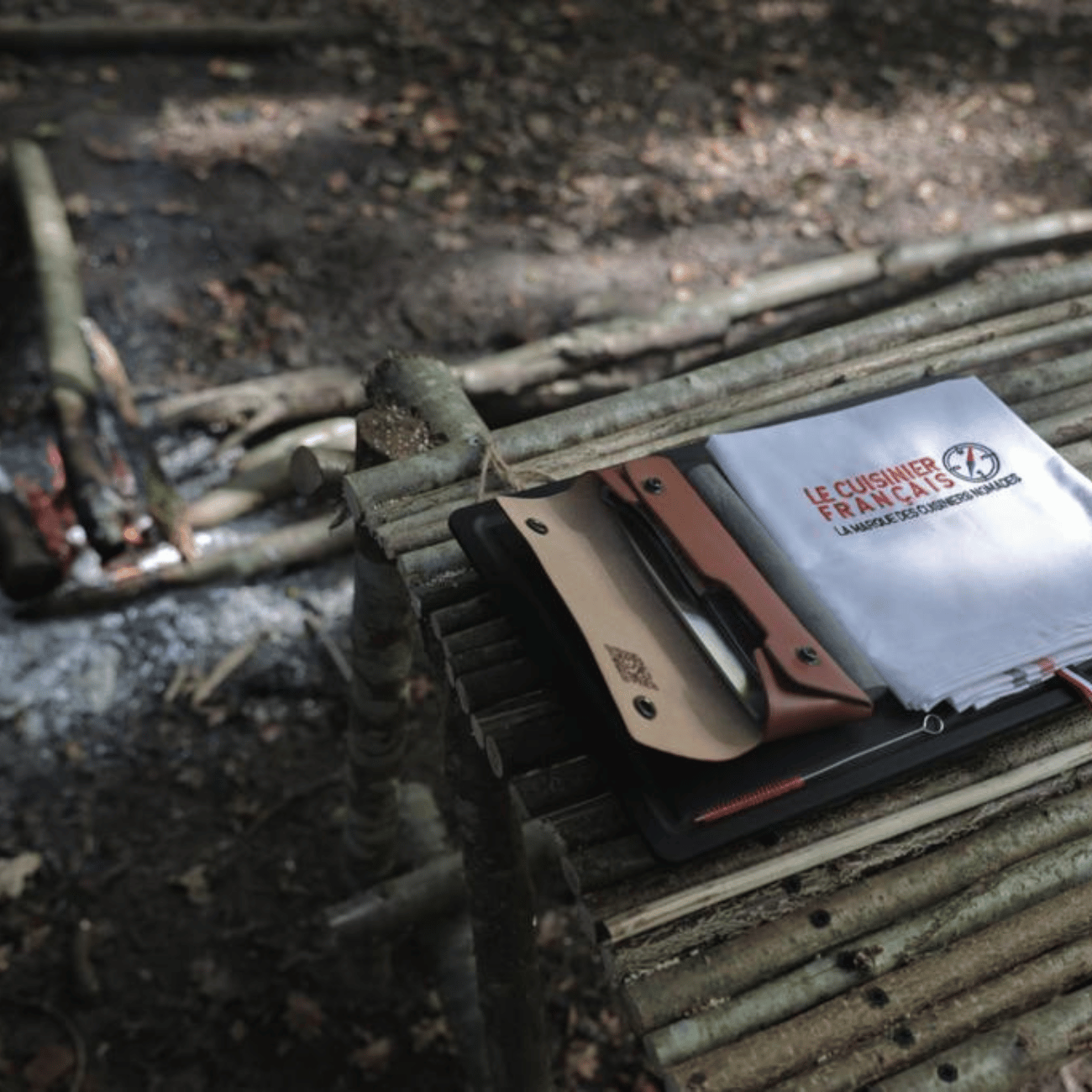 Set du cuisinier nomade en bivouac : étui en cuir ouvert et torchon Le Cuisinier Français posés sur une table en bois.